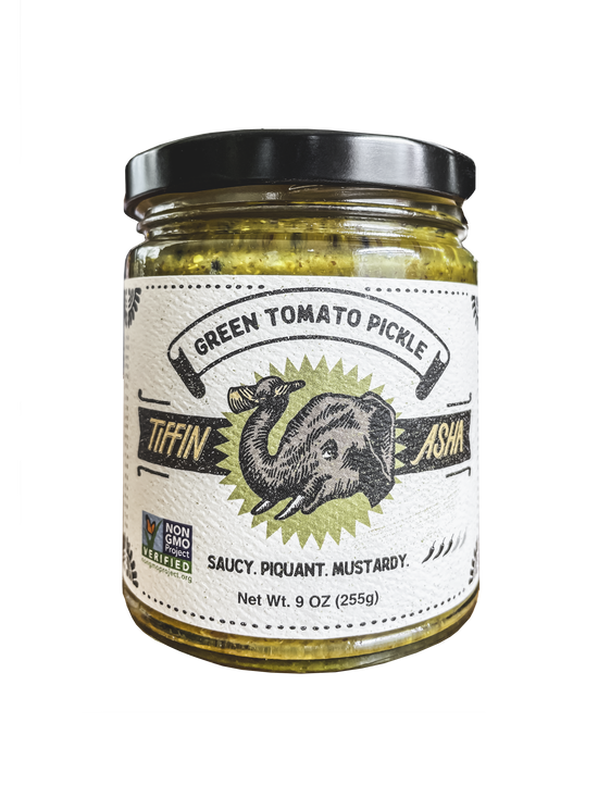 9 oz glass jar with black lid. Label reads: Tiffin Asha, with an elephant head and its trunk holding a dosa, green tomato pickle, NON GMO project VERIFIED, tasting notes read: saucy, piquant, mustardy, shows 3 chili peppers for heat level.