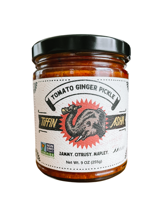 9 oz glass jar with black lid. Label reads: Tiffin Asha, with an elephant head and its trunk holding a dosa, Tomato Ginger Pickle, NON GMO project VERIFIED, tasting notes read: jammy, citrusy, mapley, shows 2 chili peppers for heat level.